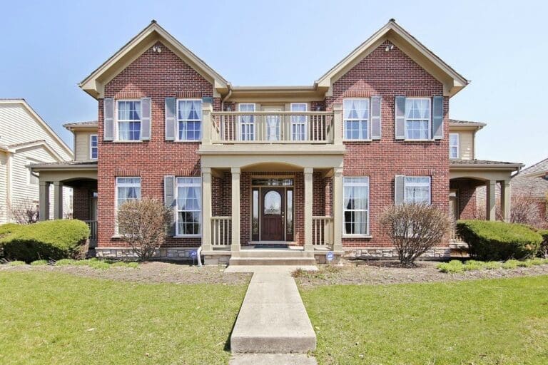 A two-story red brick house with multiple windows, tan trim, and a central front porch welcomes you. Neatly trimmed bushes and a manicured lawn complete the inviting exterior—perfect for a memorable home tour.