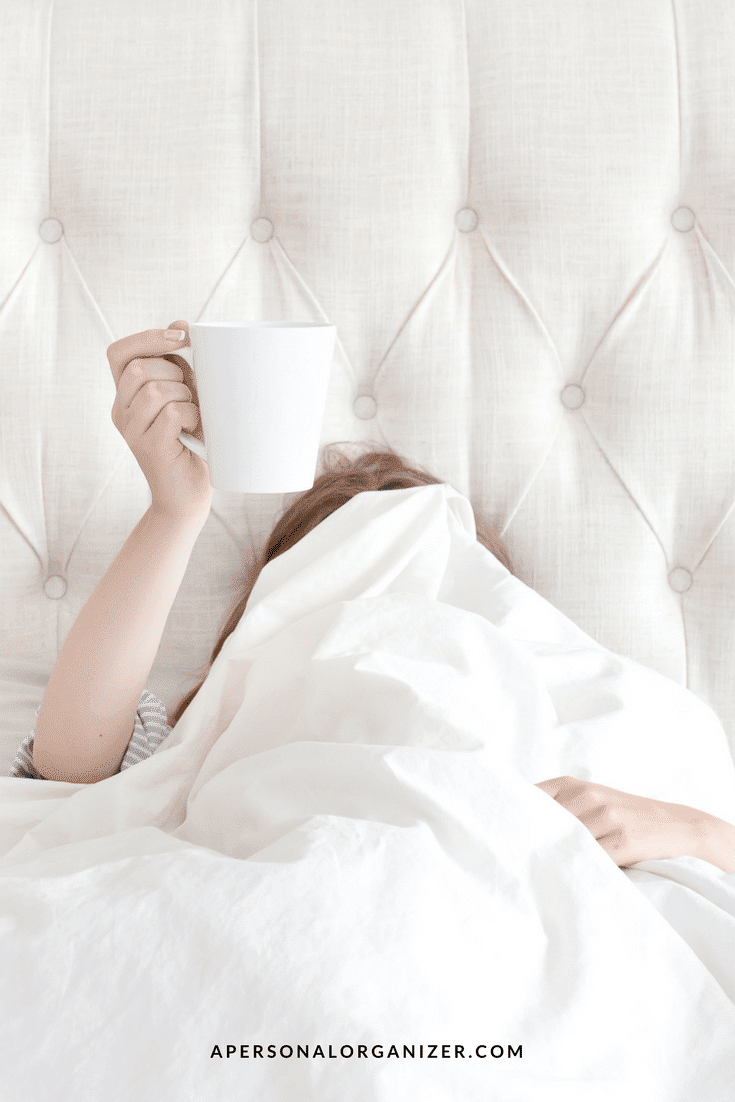 A person lying in bed is covered with a white blanket, with only one arm and the top of their head visible. They are holding up a white mug, perhaps enjoying their morning routine. The headboard is tufted and light-colored. The website "APERSONALORGANIZER.COM" is at the bottom.
