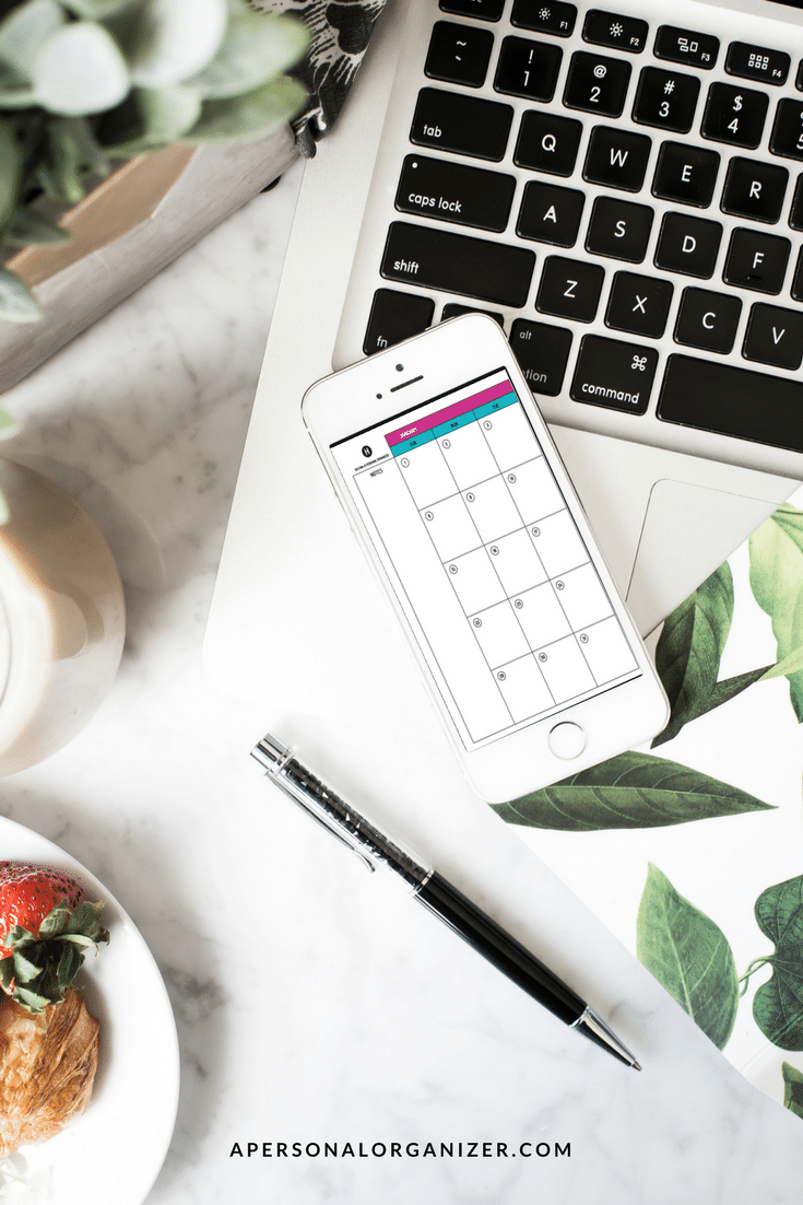 A tidy workspace featuring a smartphone displaying a calendar app, placed on a laptop keyboard. Nearby are a pen, a decorative notebook with green leaf designs, a potted plant, and a plate with strawberries and a pastry. This serene setup doubles as an effective organizing tool; visit "apersonalorganizer.com" for more tips.