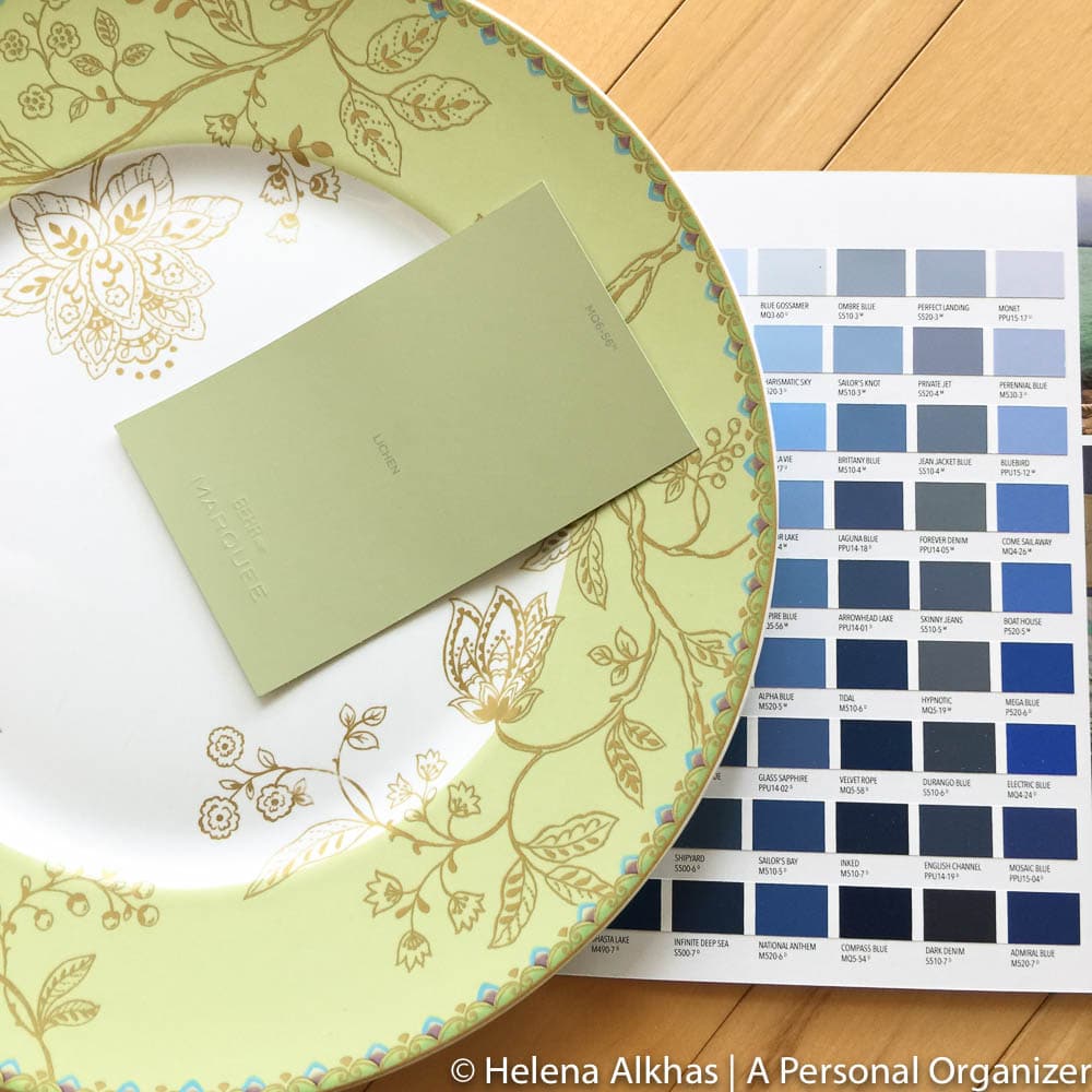 A large decorative plate with a floral pattern holds a green paint sample card labeled "Lounge," perfect for choosing exterior colors. A color swatch booklet with various shades of blue and neutral home colors lies beside the plate. Image credit: Helena Alkhas at A Personal Organizer.