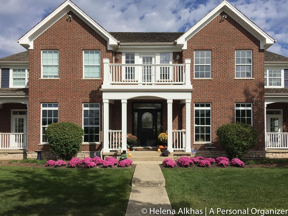 A large two-story brick house with white trim and multiple windows. There is a central entrance with white columns, a balcony above it, and a concrete pathway leading to the front door. The lawn is well-maintained with pink flowers and bushes on either side of the path, beautifully complementing the home's exterior colors.