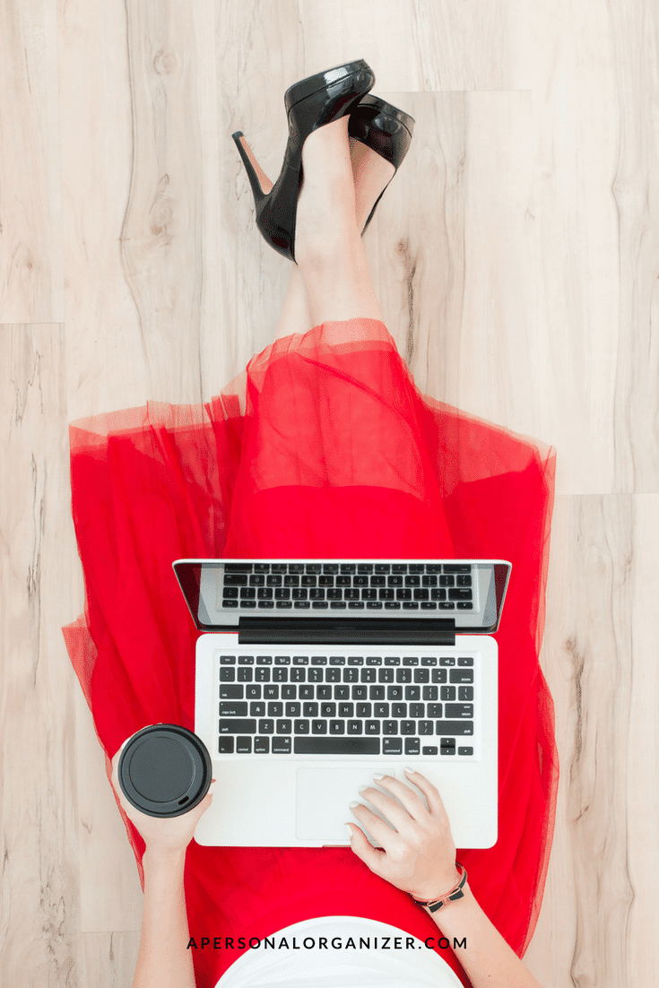 A top-down view of a person wearing a red dress and black high heels, sitting on a light wood floor while working on a silver laptop. The person holds a black cup in their left hand. The website address "APERSONALORGANIZER.COM" is at the bottom of the image, creating a Pinterest-perfect scene.