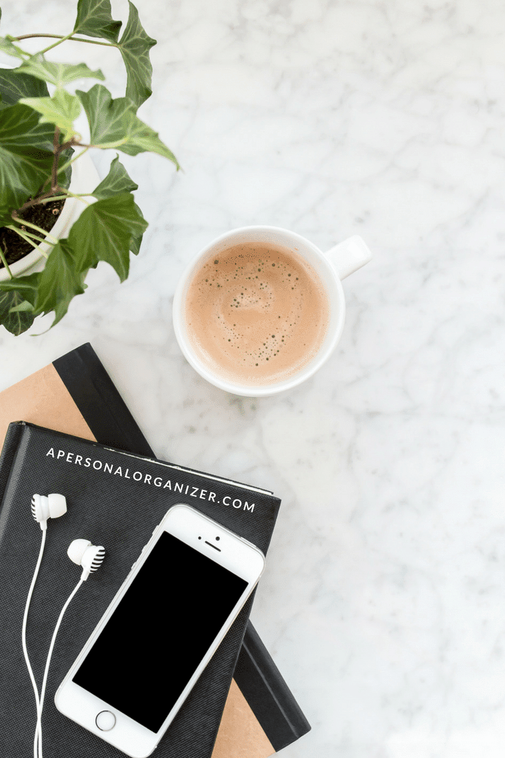 A marble surface with a mug of coffee, a plant, a pair of earphones, a smartphone, and two stacked notebooks—one adorned with "apersonalorganizer.com" on the cover. Perfectly Pinterest-worthy setup for productivity!