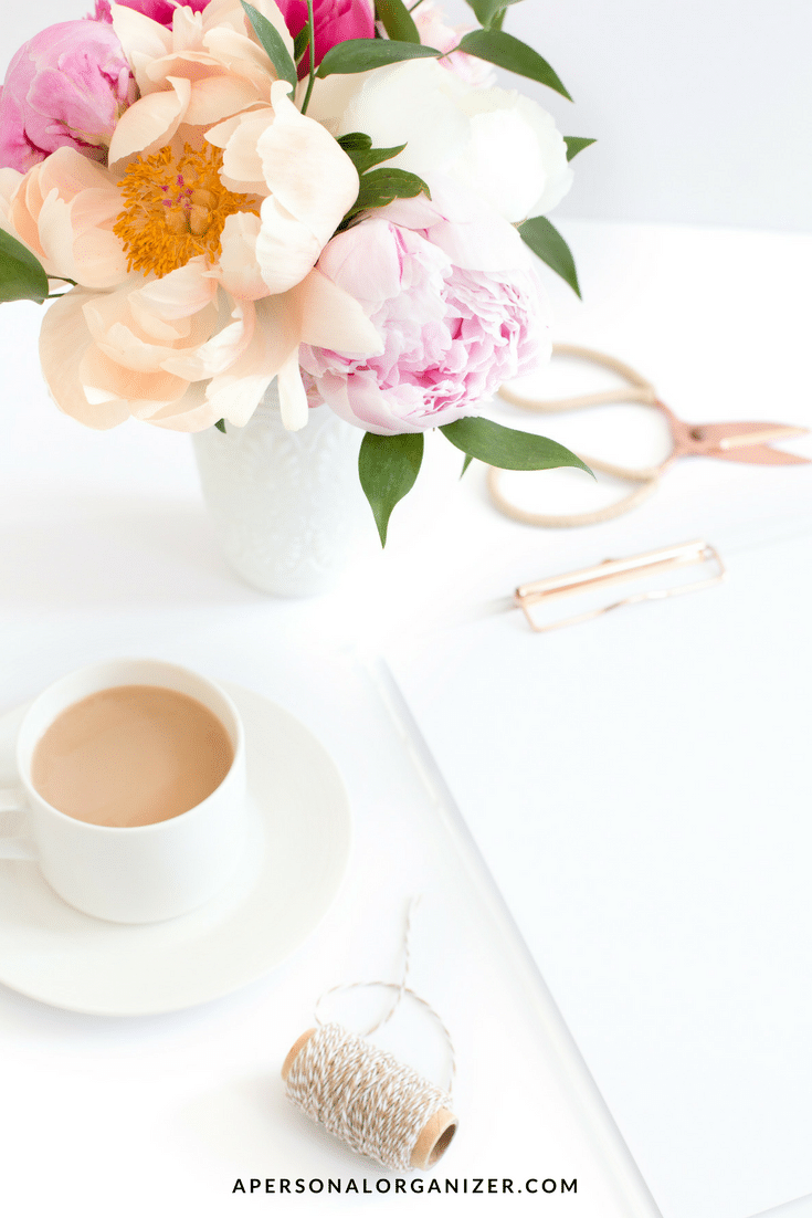A stylish desk setup designed to keep you productive at work features a white cup of coffee, a bouquet of pink and white flowers in a vase, a pair of gold scissors, a spool of twine, and a clipboard with white paper. The website "apersonalorganizer.com" is visible at the bottom of the image.