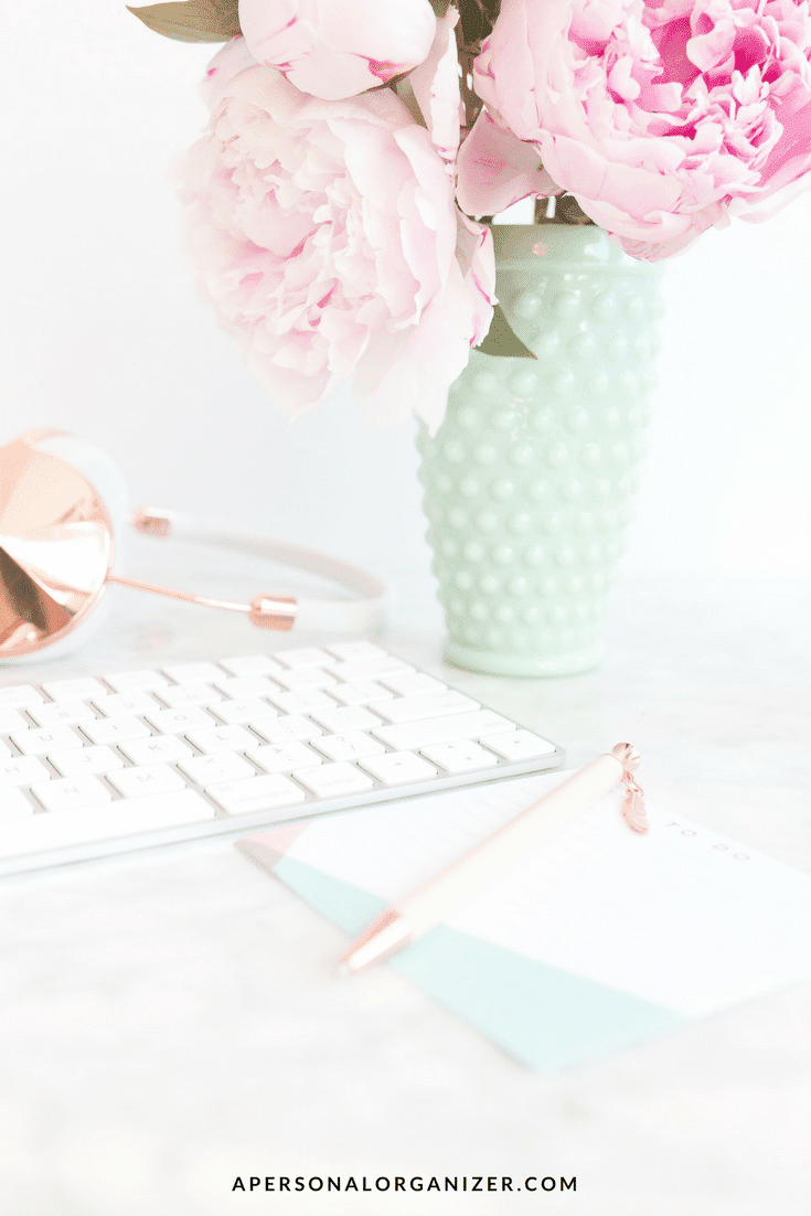 A workspace featuring a white keyboard, a notepad with a pen, a pair of headphones, and a green vase filled with pink peonies. The background is light and airy. "Stay Organized" is the focus here, showcased prominently on apersonalorganizer.com at the bottom of the image.