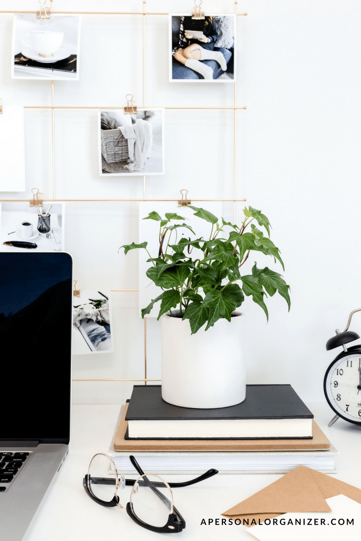 A minimalist workspace featuring a laptop, a white potted plant on stacked books, eyeglasses, a white analog clock, and a photo wire grid with black and white pictures clipped to it. The surface is clean and organized, giving a sleek and tidy appearance.