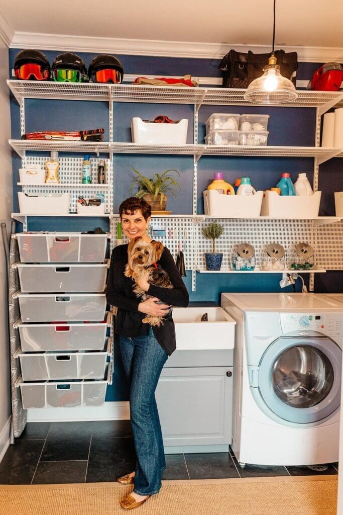 A person stands in a well-organized laundry room with blue walls, holding two small dogs. Shelves filled with various household items, including laundry supplies and storage bins, are mounted on the wall above a washing machine and a sink.
