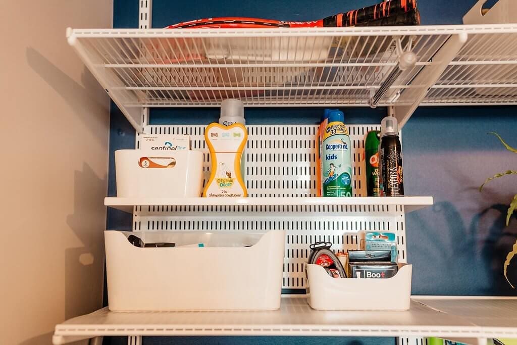 A metal shelving unit in the laundry room holds various items, including a tennis racket, bottles of sunscreen, bug spray, and two white bins with personal care products, all set against a blue wall.