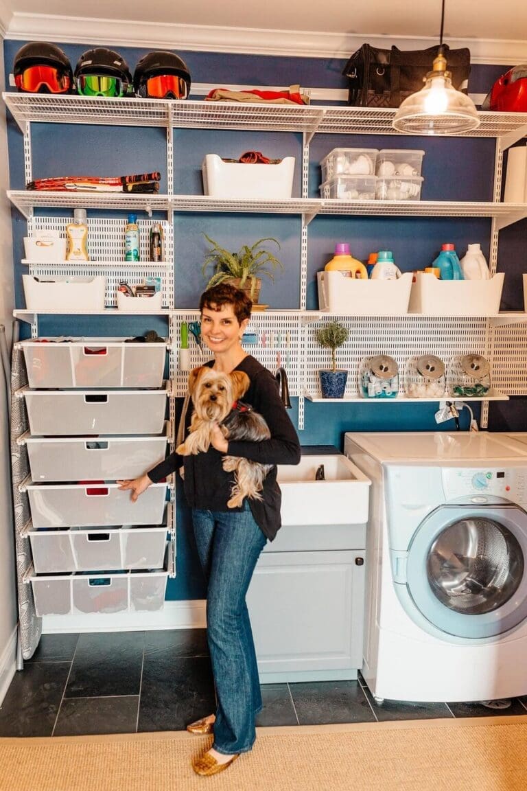 A woman holding a small dog stands in a well-organized laundry room with blue walls. Behind her are shelves stocked with cleaning supplies, helmets, and storage bins. Next to her is a white laundry sink and a front-loading washing machine.