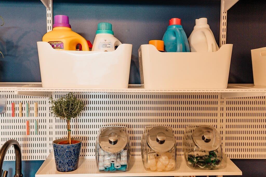 Shelves in this laundry room hold bottles of detergent in white bins, with jars below for laundry pods and dryer balls. A small potted plant sits on the lower shelf, all set against a dark blue wall.