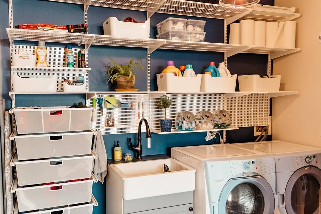 Neatly arranged laundry room with shelves for cleaning supplies, baskets, and plants above a sink. Drawers sit below the sink, with a washing machine and dryer beside it. A blue accent wall completes this functional laundry room setup.