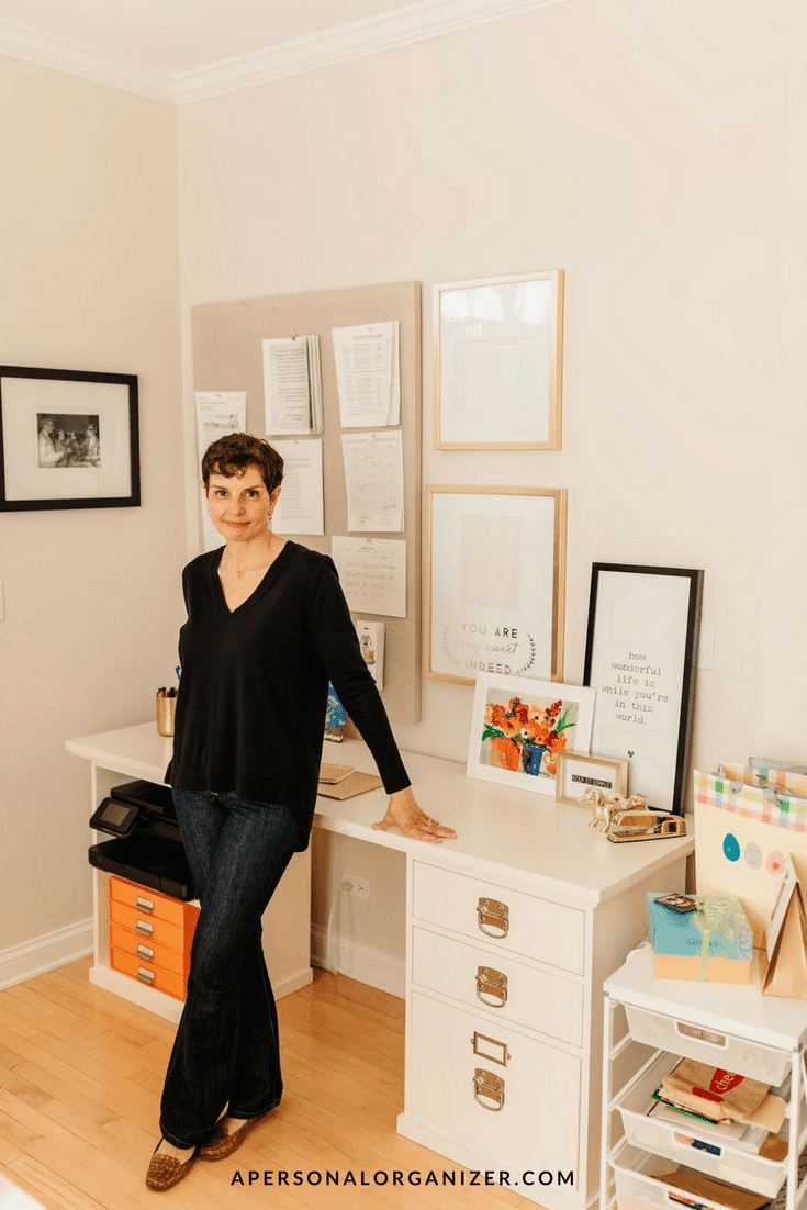 A woman in a black long-sleeve top and jeans stands next to a white desk in a well-organized home office. The desk has office supplies, paperwork, and framed prints. The room features white walls and wood flooring, creating an ideal space for her organizing business.