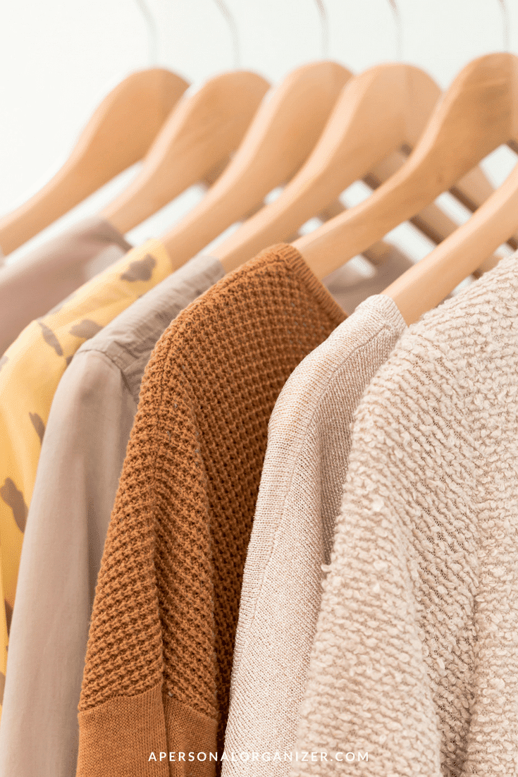 A close-up view of wooden hangers holding various textured sweaters and tops in neutral and earthy tones, including beige, brown, and a yellow patterned piece. The garments are neatly organized like attire for a professional conference, creating a visually pleasing arrangement.
