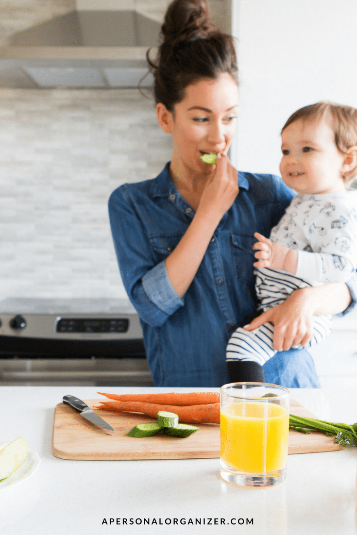 A woman stands in a kitchen holding a toddler, embodying work-life balance. She is eating a slice of cucumber and wearing a blue denim shirt. The toddler is in pajamas with a black and white pattern. On the counter are a glass of orange juice, a knife, carrot sticks, and cucumber slices.