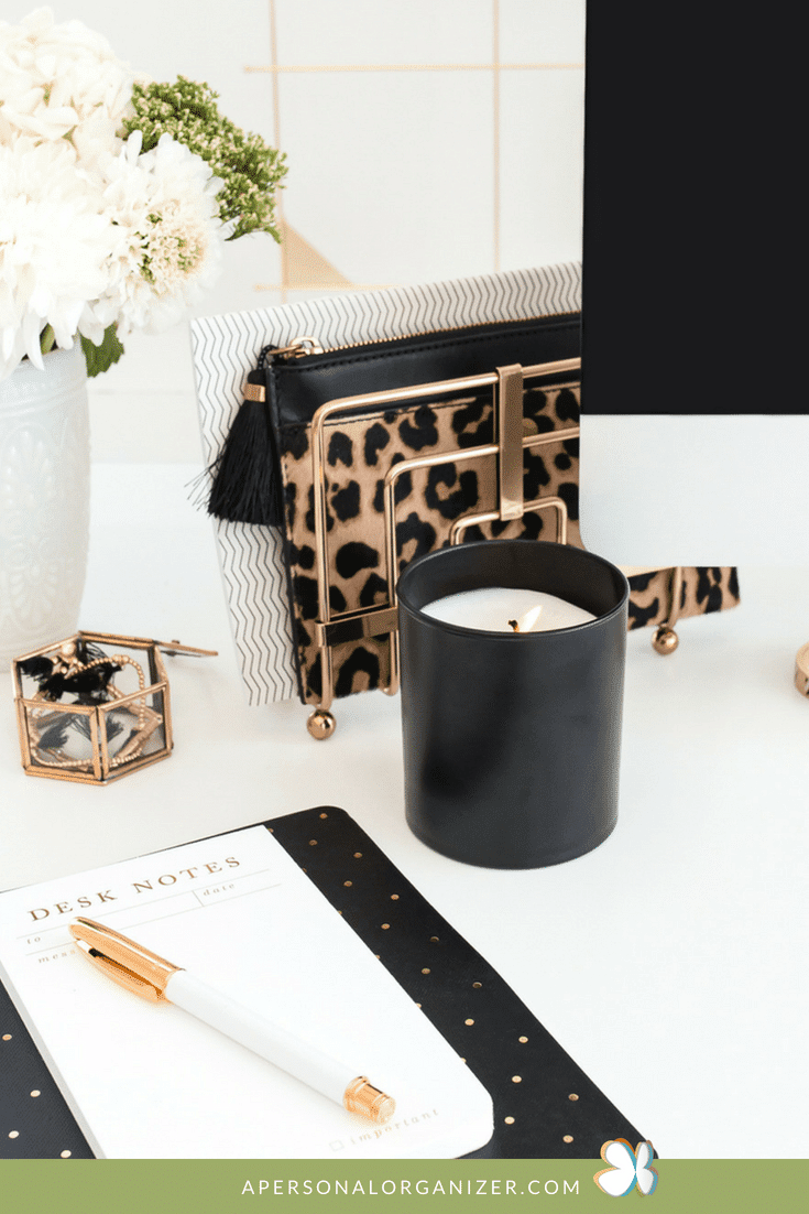 A neatly organized home office desk with a black candle, gold pen on a desk notes pad, a small gold box with paper clips, and a leopard print binder. A vase with white flowers is in the background, along with a computer monitor. Text at the bottom reads "APERSONALORGANIZER.