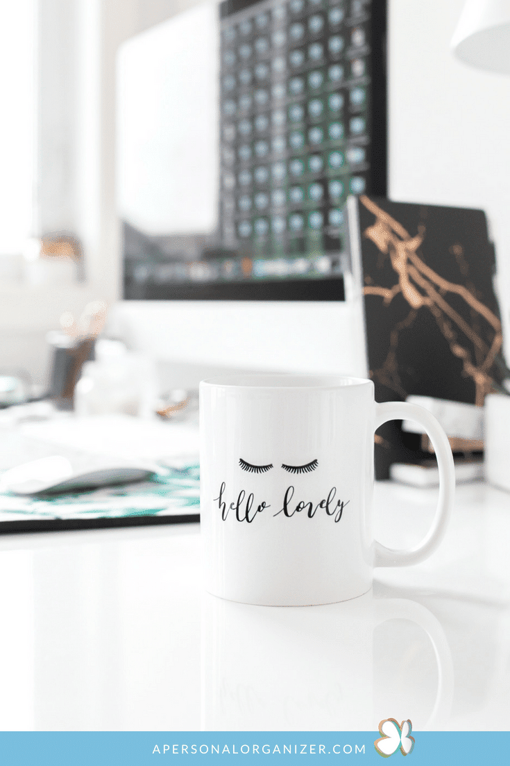 A white coffee mug with the phrase "hello lovely" and an illustration of closed eyes rests on a cluttered desk. In the background, a computer screen glows, alongside a marbled notebook and various office supplies, all contributing to a productive workspace.