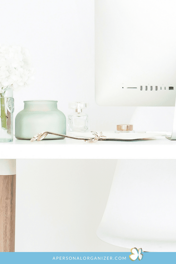 A minimalist white desk setup featuring an iMac, a small vase with white flowers, a green candle holder, a perfume bottle, and a pair of glasses. The desk has a wooden leg and a white chair. The scene is bright and clean, with neatly organizing the cords beneath ensuring no clutter, and website text at the bottom.