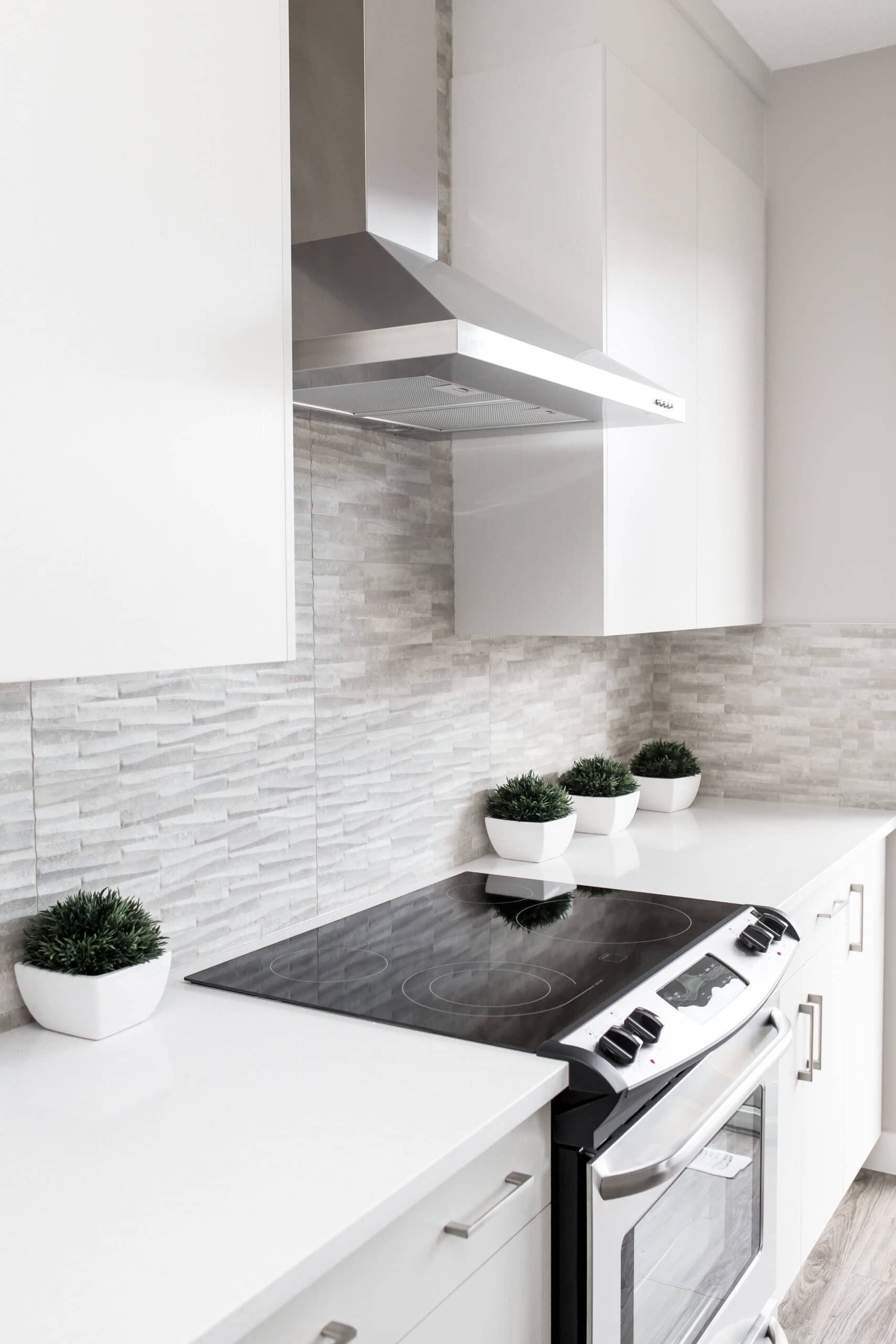 A modern kitchen with white cabinetry, stainless steel stove and hood, and a sleek black cooktop. The backsplash features light grey textured tiles, making the space easy to incorporate into your cleaning routine. Four small potted plants in white square pots are neatly lined up on the counter.