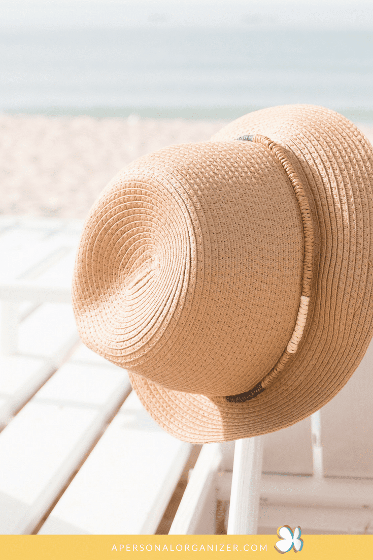 A beige straw hat hanging on a white wooden chair, with a sandy beach and the sea in the background. The sun is shining, casting soft shadows, creating a serene and relaxed atmosphere—reminding you of things to take to the beach. A yellow strip at the bottom displays a website address: apersonalorganizer.com.