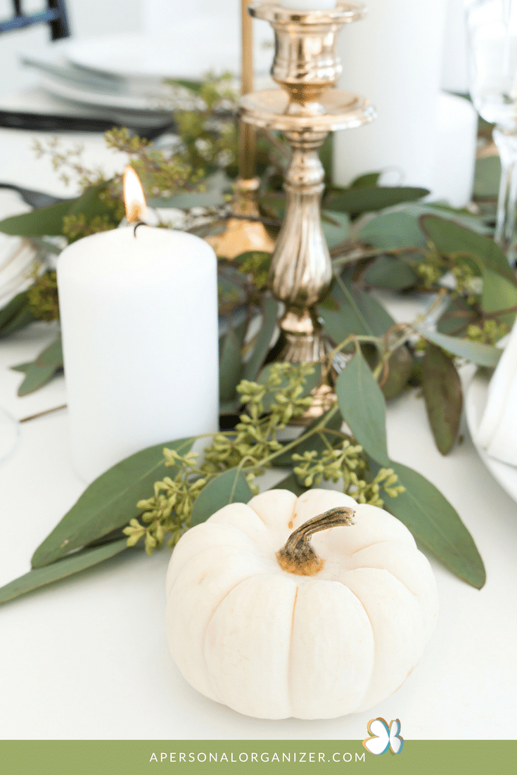A white pumpkin is placed on a decorated table with greenery, a lit white pillar candle, and a silver candlestick. The scene, devoid of clutter, suggests a festive, autumnal or Thanksgiving setting. A website URL "apersonalorganizer.com" is visible at the bottom of the image.