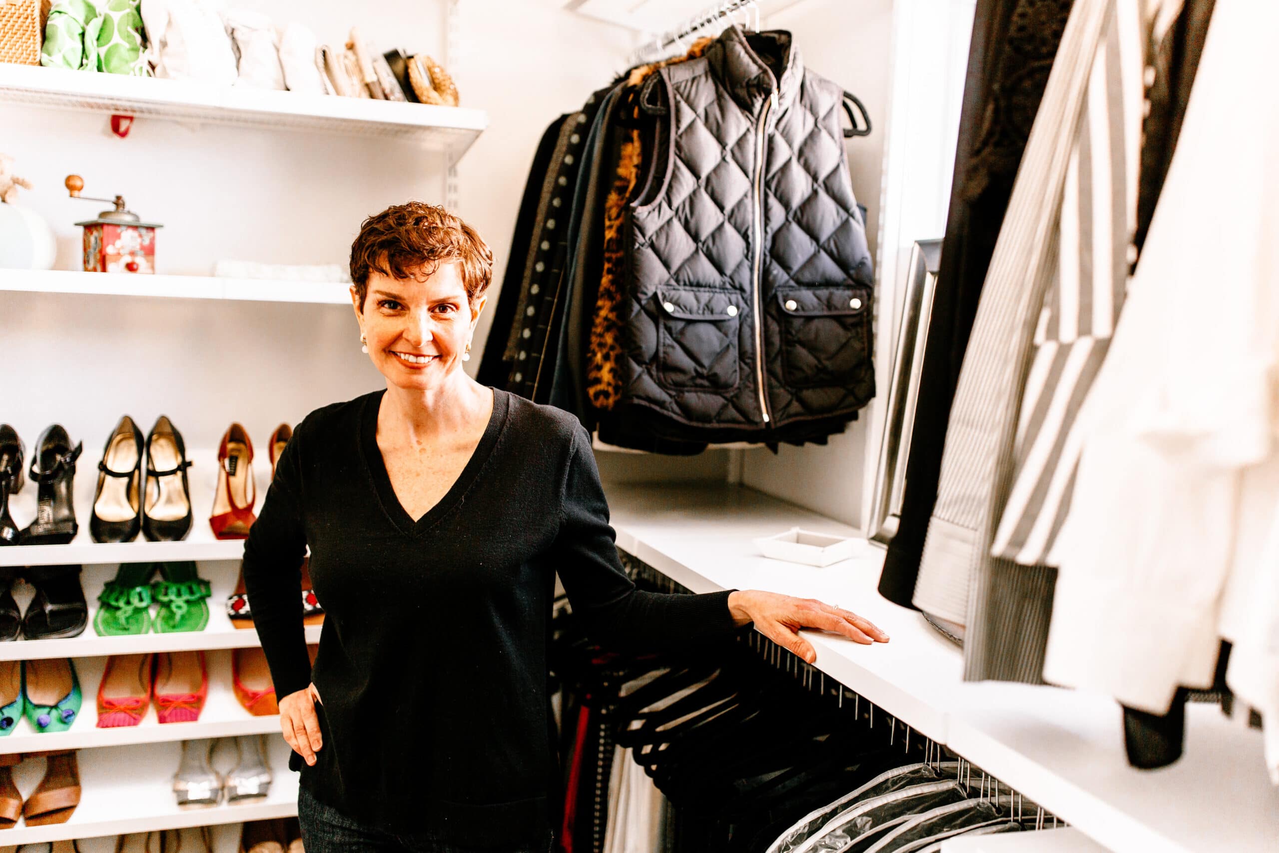 A woman with short hair and a black top stands in a well-organized closet, showcasing expert closet organizing. The space features neatly arranged shoes, clothes on hangers, and a shelf with folded items. She smiles, resting one hand on the shelf.