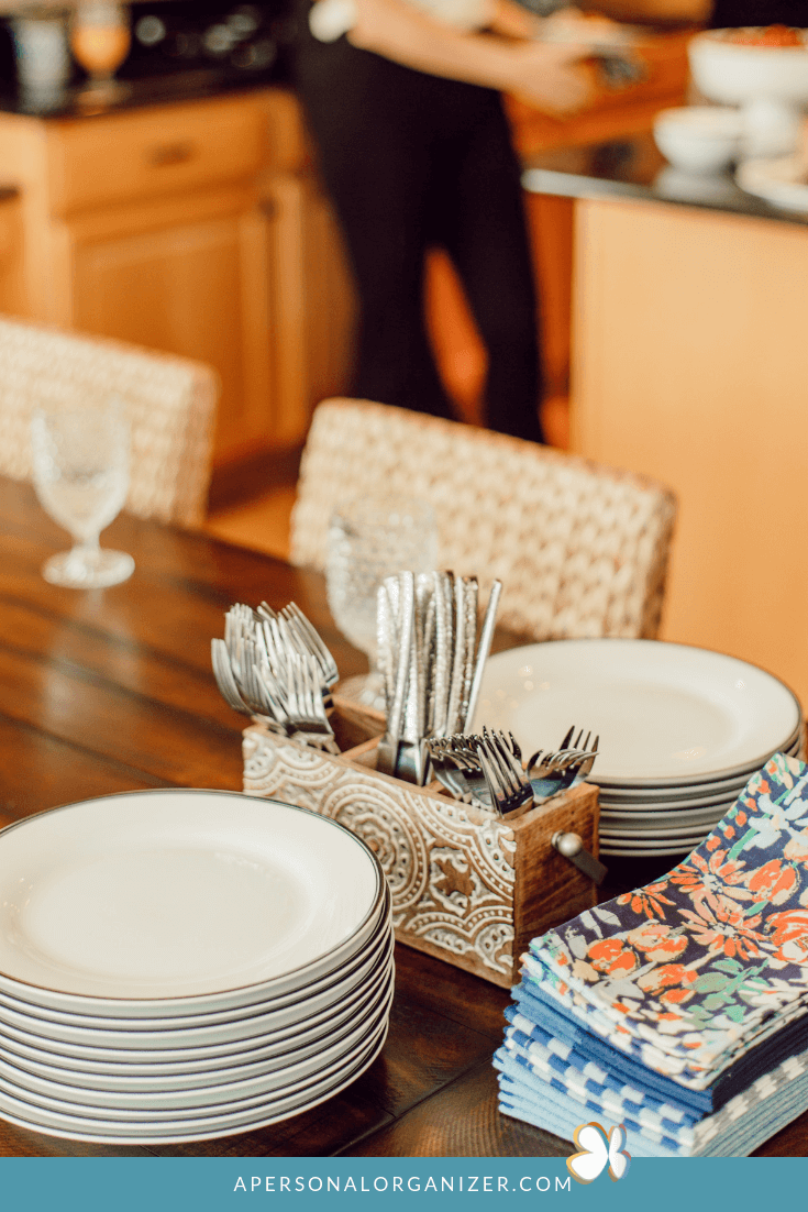 A dining table is set for brunch with neatly stacked white plates, a holder with arranged cutlery, and a pile of folded, colorful napkins. A blurred person can be seen in the background, working in the kitchen. The table is situated in a cozy wooden-themed kitchen area.
