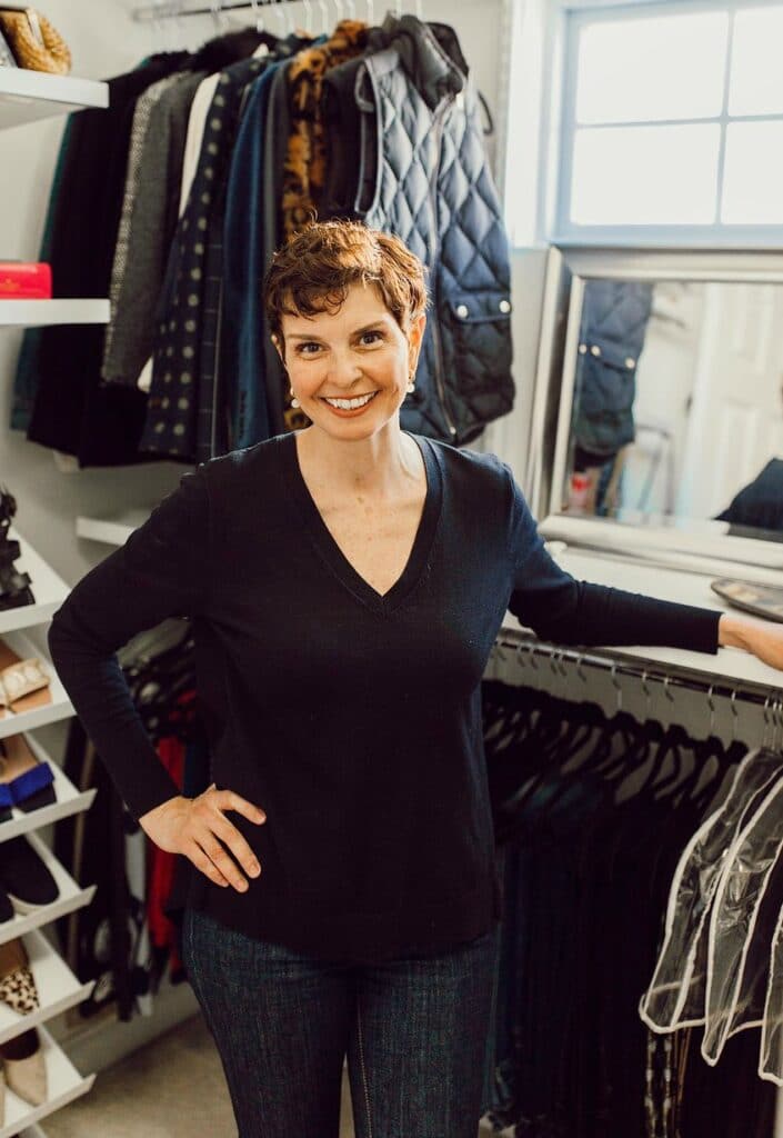 A smiling woman with short curly hair stands in a walk-in closet. She wears a black V-neck top and jeans. Behind her, shelves filled with clothes, shoes, and accessories are meticulously arranged by the professional organizer. A window is visible in the background, along with a mirror reflecting part of the room.