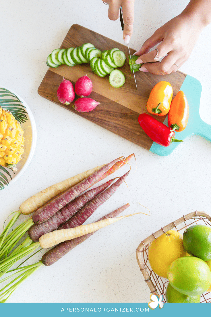 A person slices cucumbers on a cutting board with red and yellow bell peppers, showing efficient meal prep. Nearby are radishes, a bowl of pineapples, multicolored carrots, and a basket with limes and other fruits. "APERSONALORGANIZER.COM" is written at the bottom.