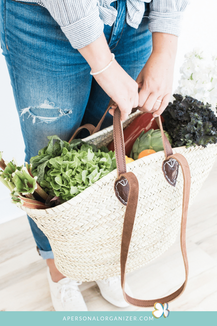 A person in a blue and white striped shirt and ripped jeans is holding a woven basket filled with fresh greens and vegetables, perfect for meal planning. The basket has brown leather handles. A logo and URL "apersonalorganizer.com" are visible at the bottom of the image.
