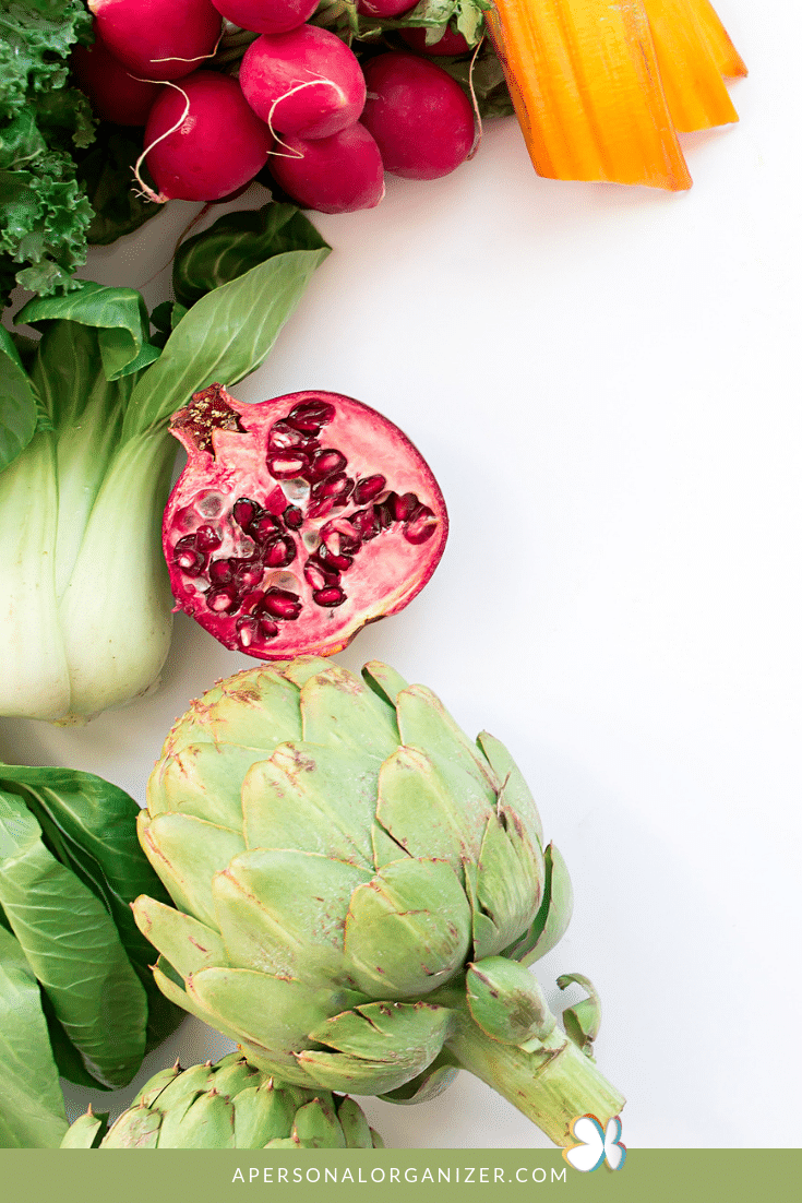 A beautifully arranged selection of fresh vegetables and fruits including artichoke, bok choy, kale, radishes, a halved pomegranate, and orange carrot sticks, displayed on a white surface. Perfect for meal planning enthusiasts, the image has a clean aesthetic with an "apersonalorganizer.com" watermark.