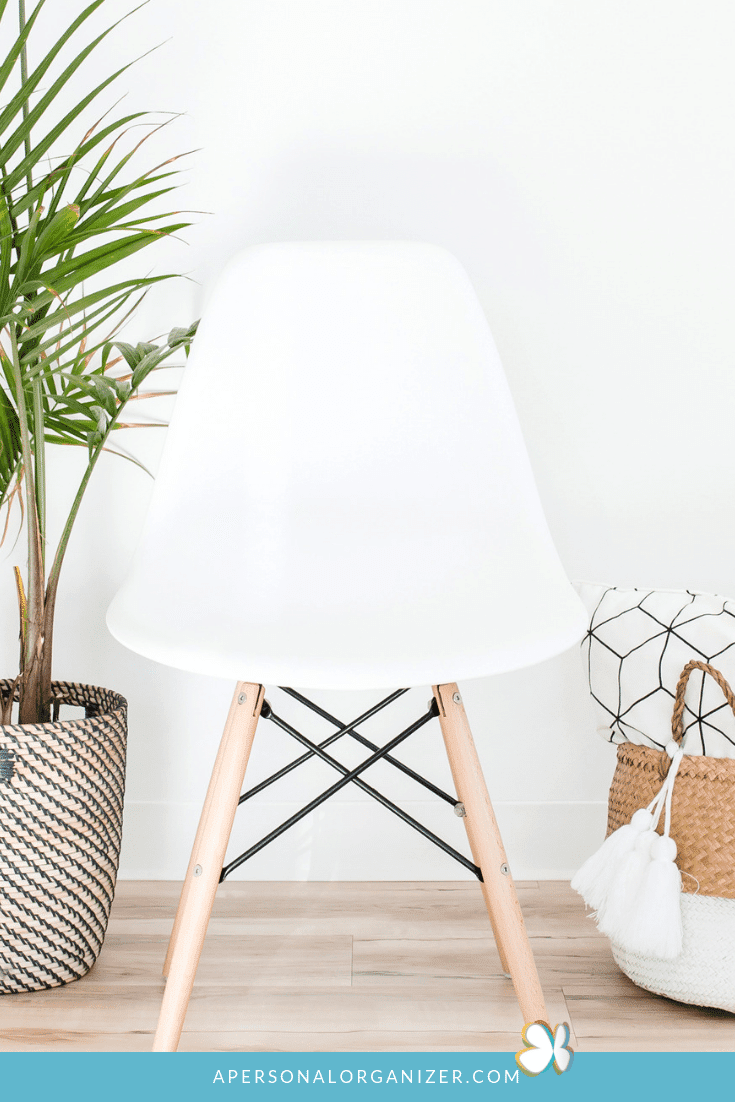 A modern white chair with wooden legs is placed against a white wall. To the left of the chair, a potted plant in a woven basket adds life, while two more woven baskets on the floor to the right maintain an organized look, free from clutter. The image boasts a clean and minimalist aesthetic.