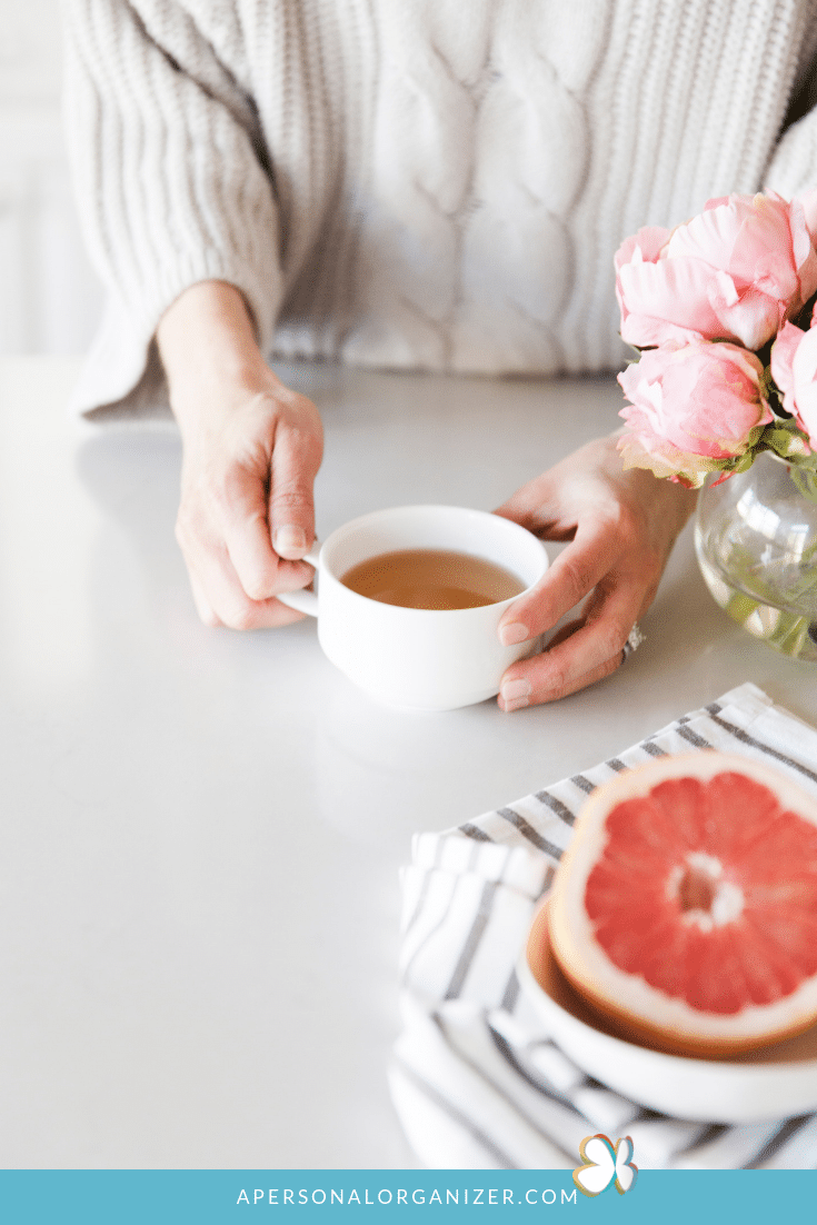 A person in a cozy sweater holds a white cup of tea on a tidy white countertop. Nearby, there is a vase with pink flowers, a folded striped cloth, and half a grapefruit. The image has a calm and inviting atmosphere, offering subtle decluttering tips for creating serene spaces.