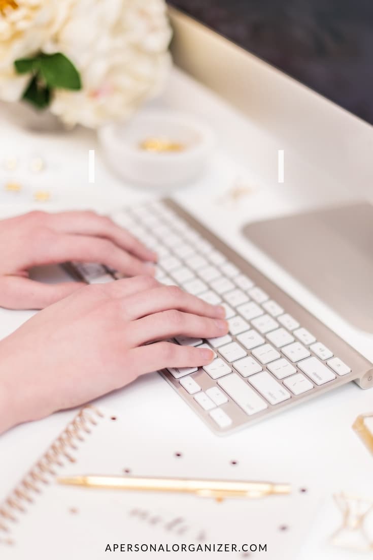 Close-up of hands typing on a white keyboard. The workspace, perfect for a business blog, includes a white desk, a gold pen, a notebook with gold accents, small decorative items, and a bouquet of white flowers in the background. The monitor is partially visible on the right side.