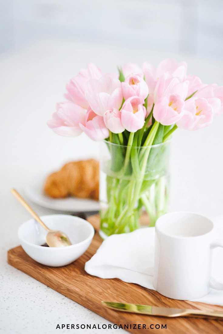 A wooden tray holds a white mug, a bowl with a spoon, and a croissant in the background. A vase filled with pink tulips is placed centrally on the tray, embodying spring organizing tips for your home. The web address apersonalorganizer.com is visible at the bottom.