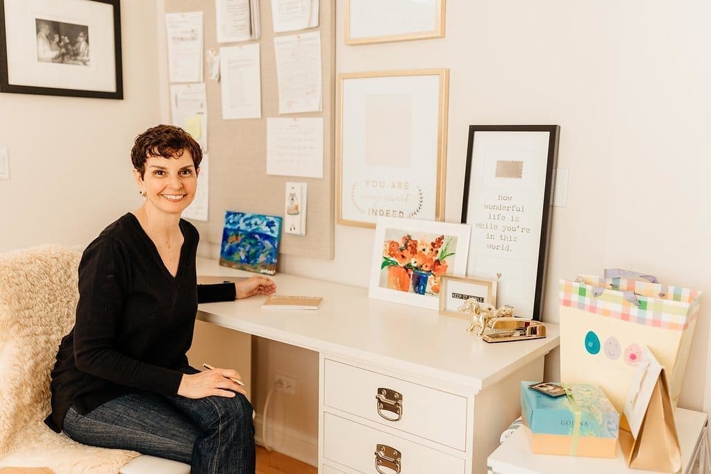 A person with short hair sits at a white desk in a cozy, well-lit home office. They are smiling and holding a notebook. The desk is adorned with colorful artwork, framed pictures, and various stationery items—a perfect setting for a professional organizer. The walls showcase more framed prints and notes.