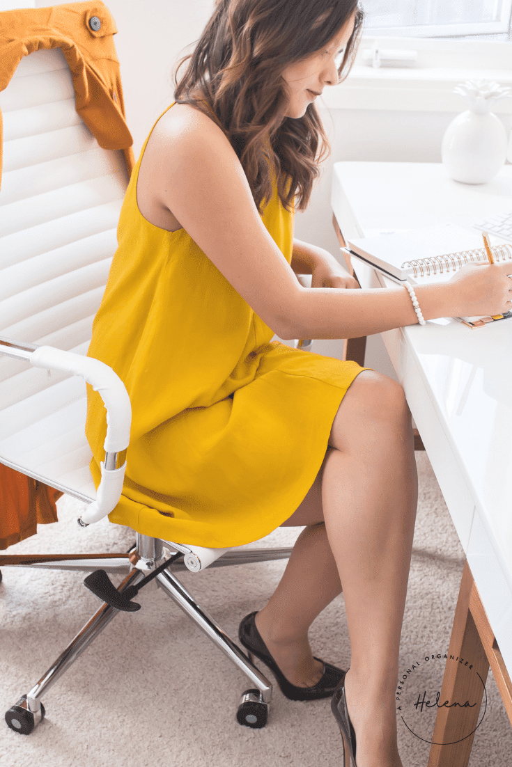 A woman in a yellow dress sits on a white chair at a desk, organizing her home office as she writes in a notebook. She has shoulder-length brown hair, wears black shoes, and the bright room features a window in the background, perfect for her work-from-home setup.