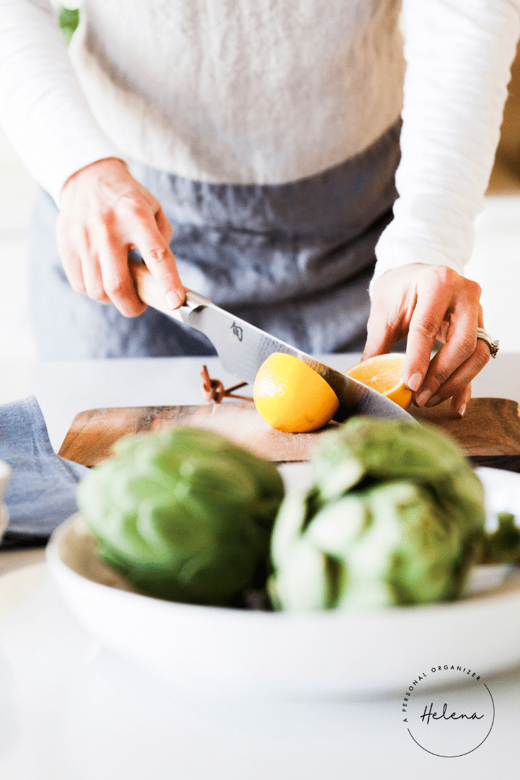 A person, embodying everyday organizing solutions, slices a lemon on a wooden cutting board. Artichokes sit elegantly on a white plate in the foreground. Dressed in a light-colored top and gray apron, they work efficiently under the "Helena" logo prominently displayed in the corner.