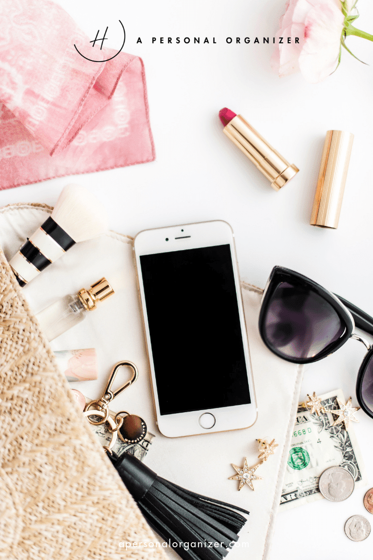 A flat lay image showcases a smartphone, sunglasses, and lipstick beside a makeup brush and star-shaped earrings on a white surface. Amid the organized commitments of fashion are a pink scarf, straw bag, gold keychain, dollar bills, and a chic tassel accessory.