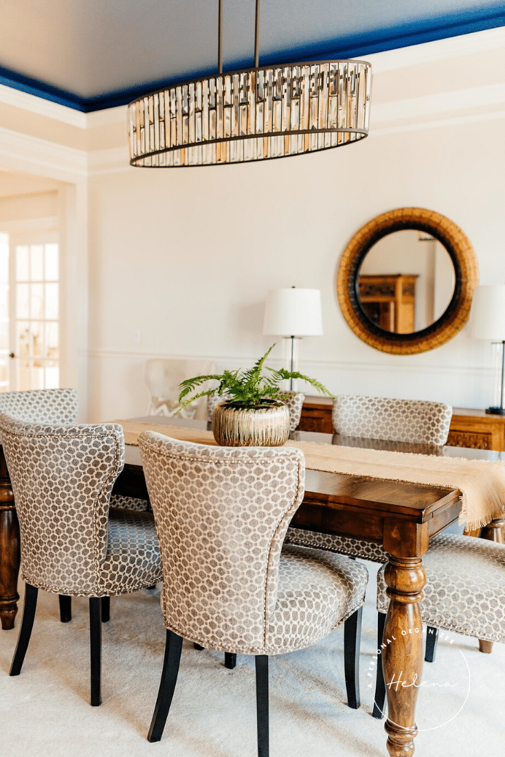 A dining room featuring a wooden table with six patterned chairs is impeccably organized. A central plant decorates the table, while a modern chandelier above illuminates the space. A round mirror and two lamps enhance the background ambiance, all beneath a beautifully painted blue ceiling.