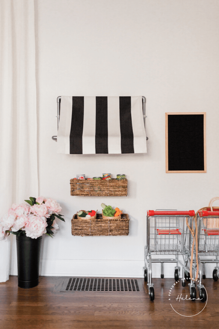 The playroom features a striped towel on a wall rack, two wicker baskets holding small grocery items, and a blank chalkboard. A vase of pink flowers sits on the floor beside two miniature shopping carts brimming with assorted items.