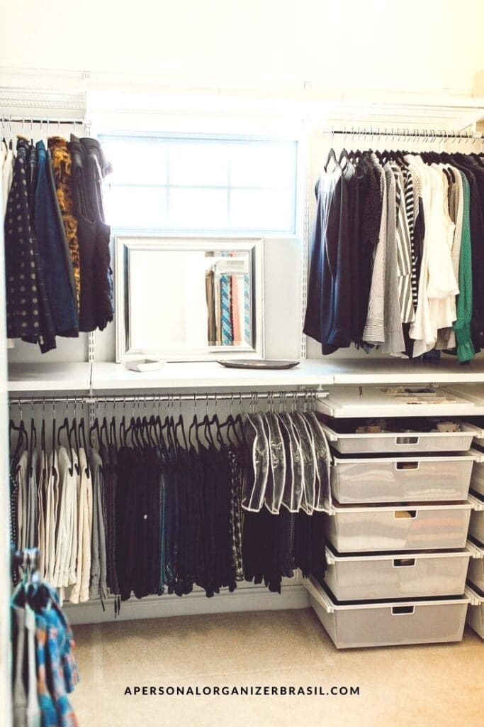 A well-organized closet, or *guarda roupa*, features shelving and hanging clothes. A mirror is positioned centrally with racks on either side. Transparent drawers store additional items, while a window bathes the space in natural light.