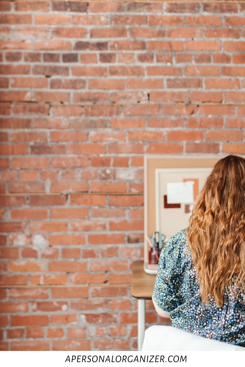 A person with long hair sits at a desk in what appears to be a casually organized college dorm room. They face a corkboard mounted on the brick wall. The website "apersonalorganizer.com" is noted at the bottom of the image, blending seamlessly into this study-friendly environment.