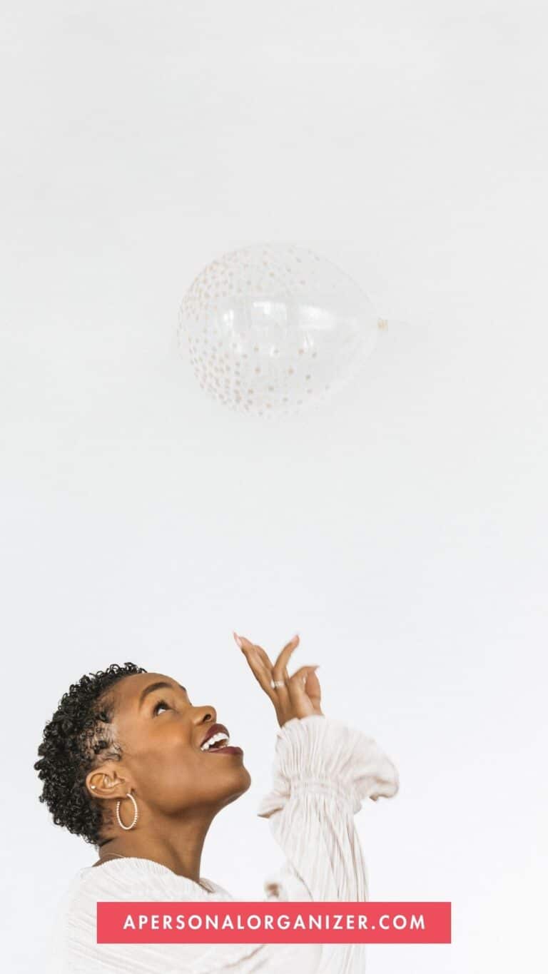 A person with short curly hair wearing a light long-sleeve top is looking up, gesturing toward a floating transparent balloon with gold dots. The plain white background adds elegance, while a red banner reads: "APERSONALORGANIZER.COM," perfect for New Year home organizing inspiration.