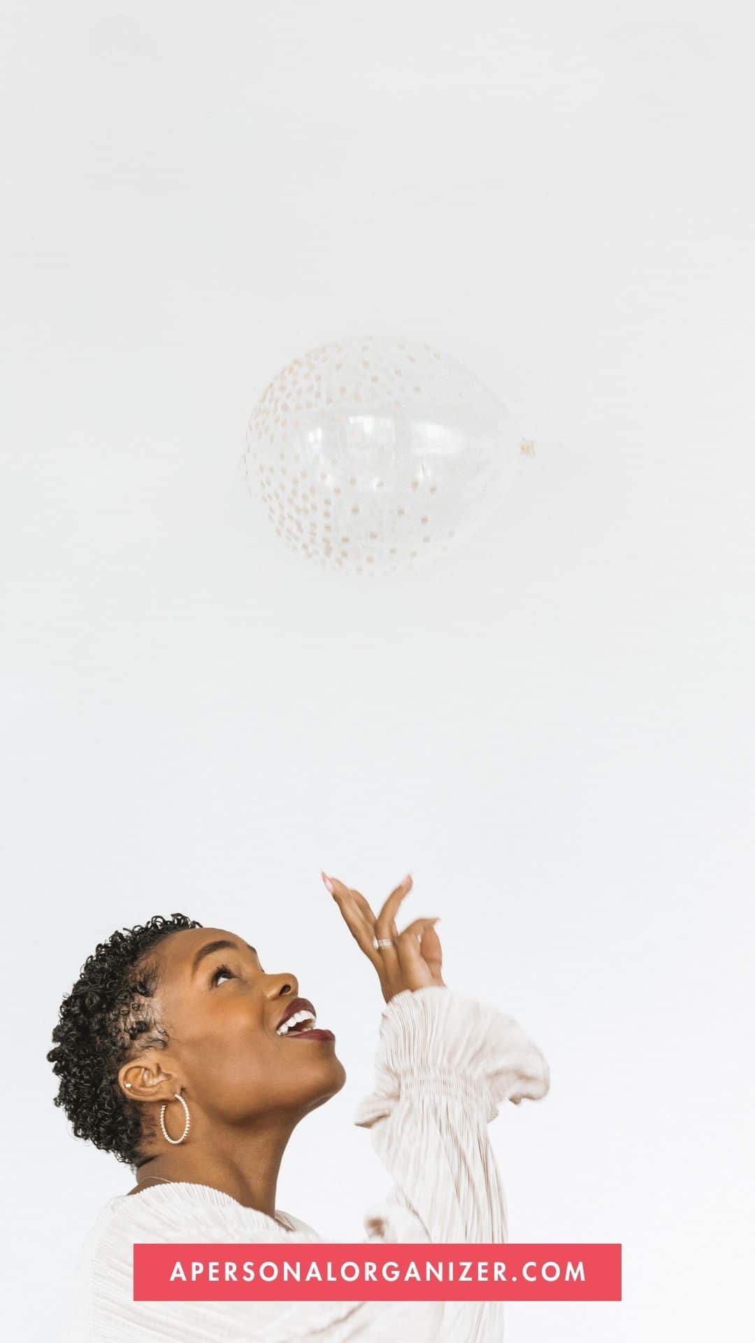 A person with short curly hair wearing a light long-sleeve top is looking up, gesturing toward a floating transparent balloon with gold dots. The plain white background adds elegance, while a red banner reads: "APERSONALORGANIZER.COM," perfect for New Year home organizing inspiration.