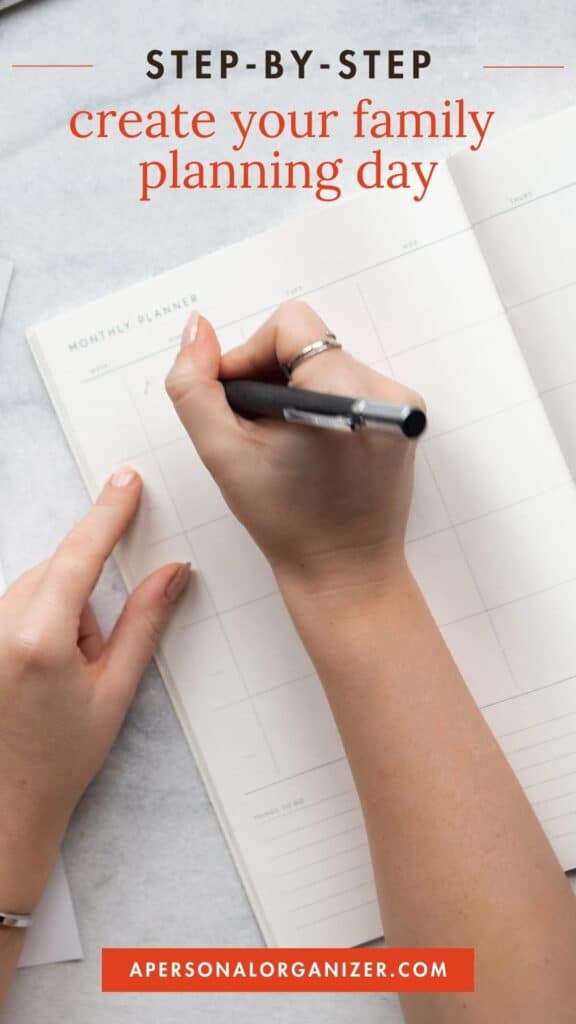 A person writes in a monthly planner on a white desk. The planner's blank squares await entries for each family planning day. Text above reads "Step-by-step create your family planning day" and below "apersonalorganizer.com.