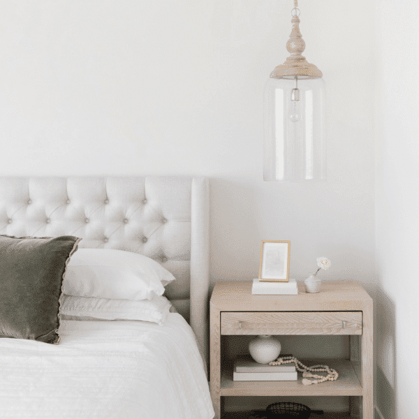 A minimalist bedroom features a gray tufted headboard and a neatly made bed with two pillows. A wooden nightstand, perfect for organizing products, holds a small framed picture, a decorative object, and a hanging glass pendant light above it against the white wall.