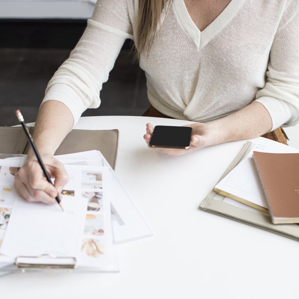 A person sits at a desk, organizing their thoughts by writing with a pencil on a notepad while holding a smartphone in the other hand. The desk is cluttered with papers, a notebook, and a folder. They are wearing a white long-sleeve shirt.