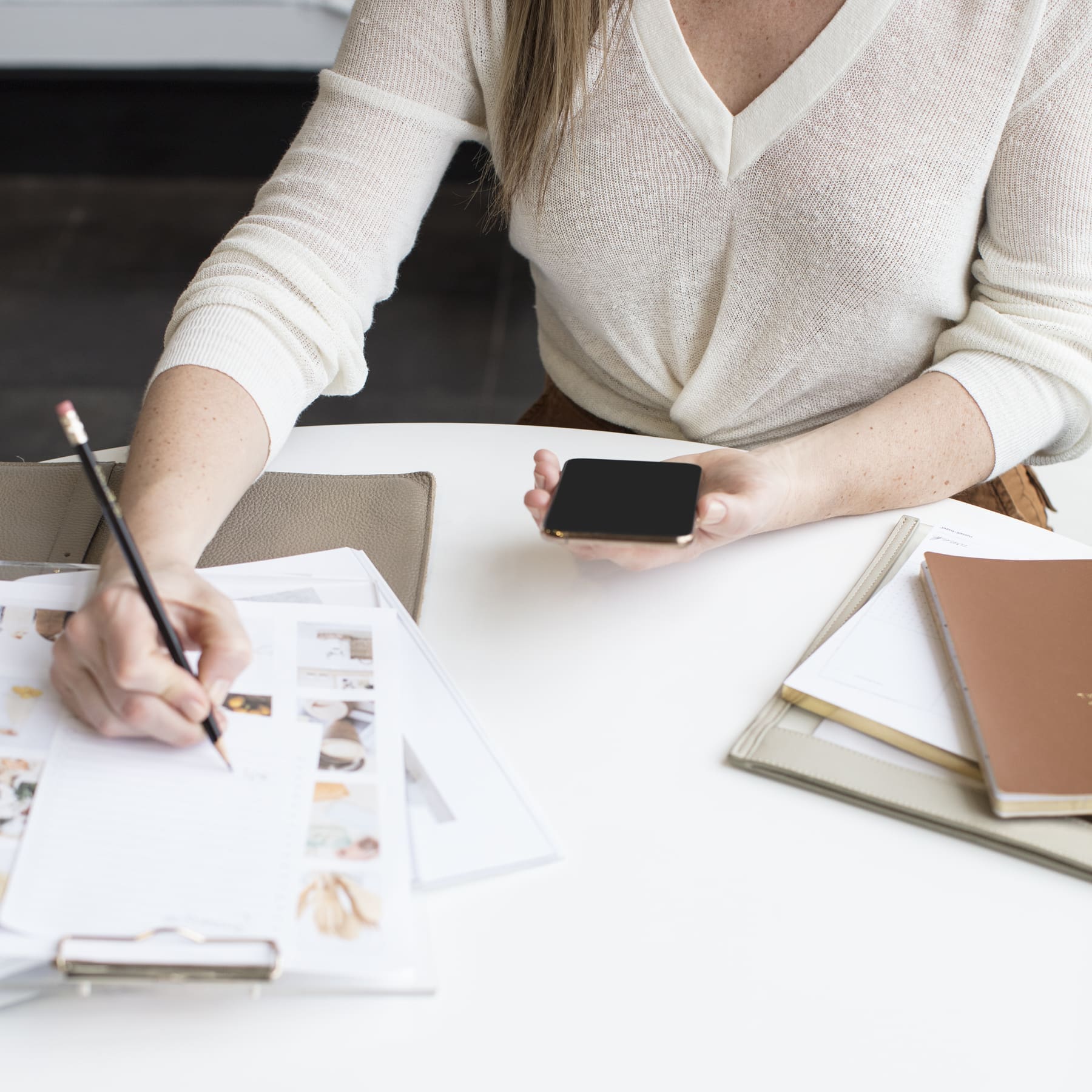 A person sits at a desk, organizing their thoughts by writing with a pencil on a notepad while holding a smartphone in the other hand. The desk is cluttered with papers, a notebook, and a folder. They are wearing a white long-sleeve shirt.