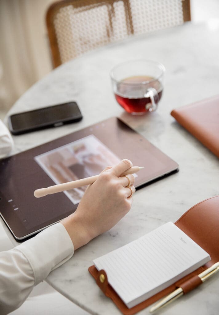 A person is engrossed in a personal organizer course, using a stylus on a tablet while seated at a round table. Nearby, a smartphone, notepad, and cup of tea rest on the marble surface, enhancing the creative ambiance.