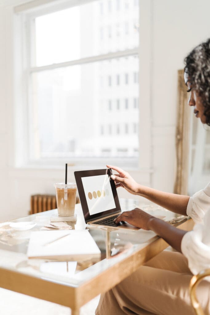 A person sits at a glass desk, using a stylus on a tablet screen displaying graphic design. Alongside iced coffee, papers detailing the latest curso personal organizer strategies are scattered nearby. A plant adds life, while large windows flood the modern office space with natural light.